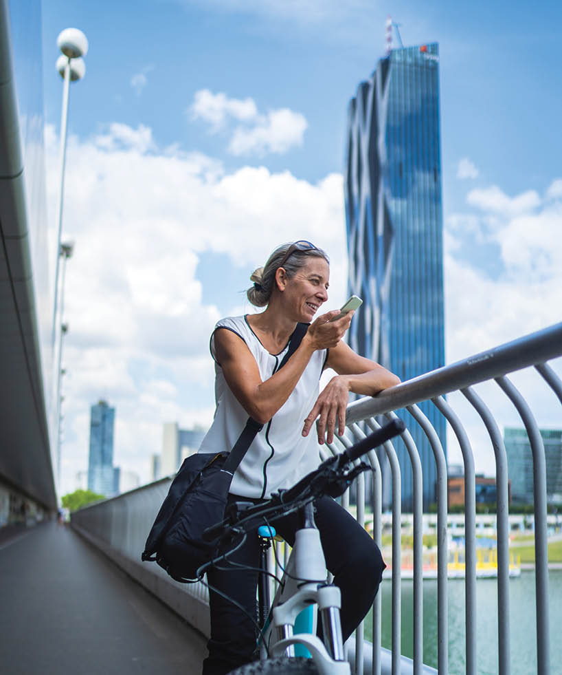 smiling 45 years old business woman on electric bicycle on bridge over river speaking message in mobile phone outdoors in european city in summer background blurred skyscraper